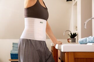 Närbild på kvinna i badrummet framför spegeln som bär Actimove Professional Line Abdominal Binder Comfort med mjukt foder
