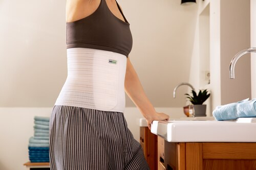 Close-up of woman in bathroom in front of the mirror, wearing an Actimove Professional Line Abdominal Binder Comfort with Soft Pad
