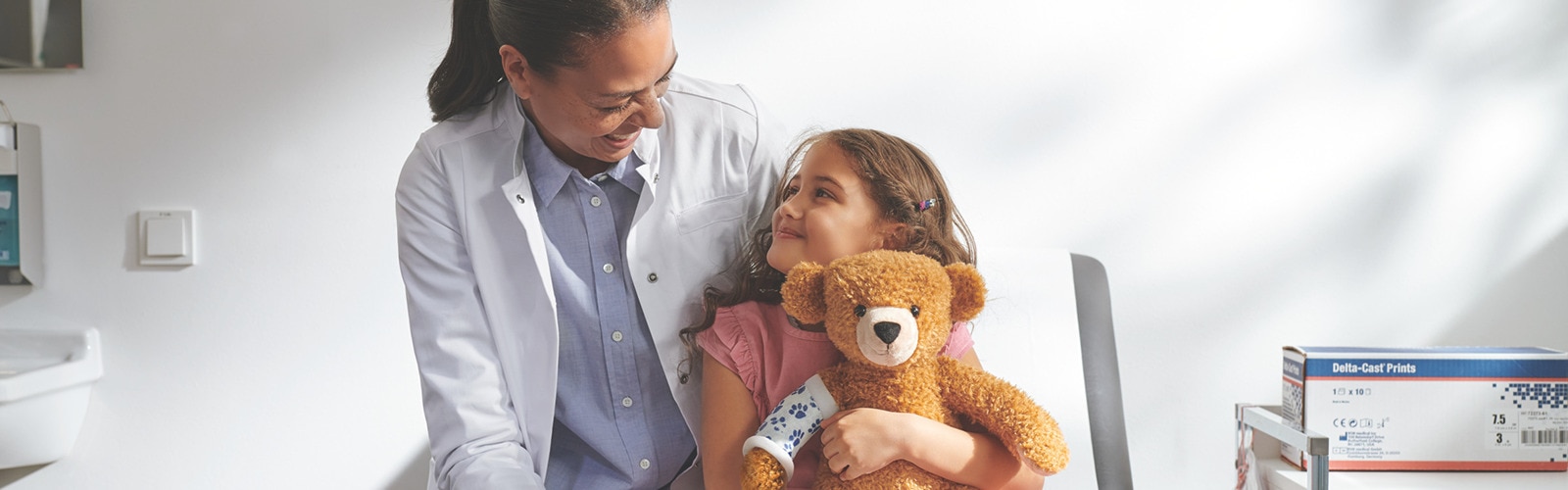 Female doctor and little girl sitting on a daybed in clinic setting, looking and smiling at each other.