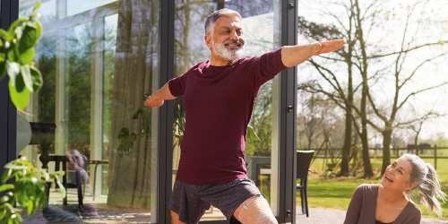 Hombre sonriente con ropa deportiva y un soporte para las rodillas hace ejercicios de estiramiento al aire libre mientras una mujer mira 