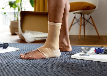 Woman standing on carpet wearing an ankle support with empty box, plant and chair in the background