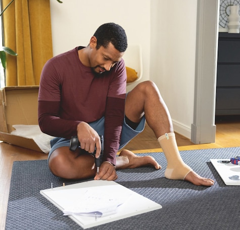 Mann som har på ankelstøtte mens han"gjør-det-selv" skrur skruer på baksiden av et kvadratisk bord Mann som har på ankelstøtte mens han"gjør-det-selv" skrur skruer på baksiden av et kvadratisk bord