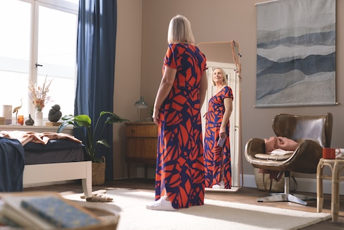 Female in a colourful dress looking at herself in a mirror in a cozy room, wearing JOBST Elvarex products.