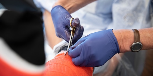 A set of hands in blue gloves holding scissors and cutting a bandage or cast from someone's arm in a clinical setting.