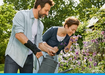 Man wearing a wrist support holds a watering can in a garden while the woman next to him smells the flowers 
