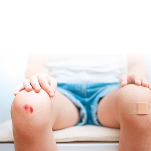 Close-up van de blote knieën van een jongen. Zijn rechterknie is geschaafd; links draagt hij een wondverband. 