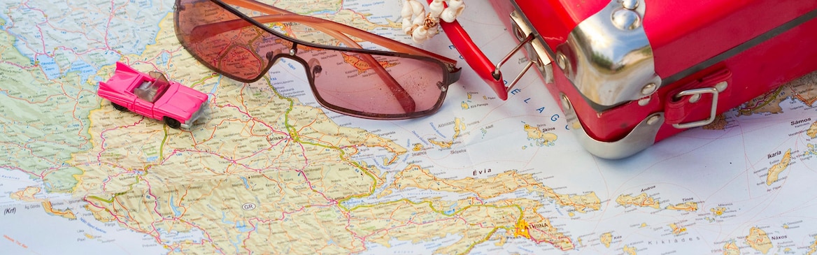 Na cestovní mapě leží: červený kufřík, sluneční brýle a růžové autíčko. 
