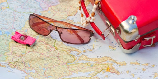 A red toy suitcase, sunglasses and a toy car are lying on a road map.