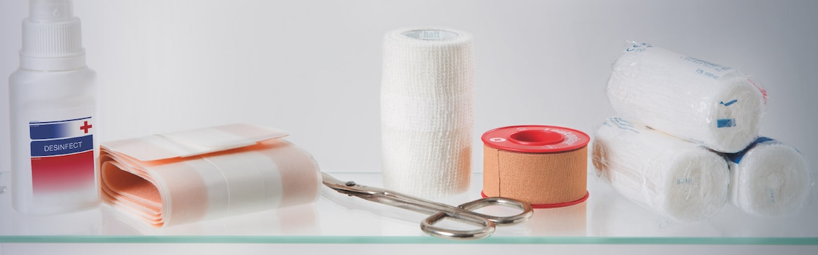 Composition of 4 rolls of bandages, a roll of adhesive wound dressing, disinfectant spray, Leukoplast fixation tape and plaster scissors on a glass table.