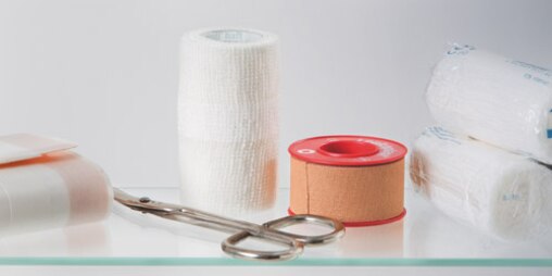 Composition of 3 gauze rolls, Leukoplast fixation tape, adhesive wound dressing and plaster scissors on a glass table.