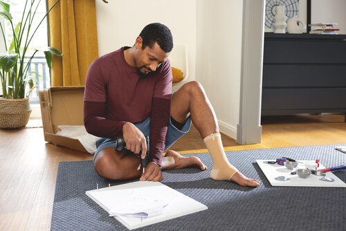 Man sitting on the floor doing home repairs, wearing a beige Actimove Arthritis Care Ankle Support
