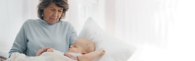 Iäkäs nainen pitelemässä nukkuvaa vauvaa Leukomed skin sensitive -laastari oikeassa käsivarressaan.