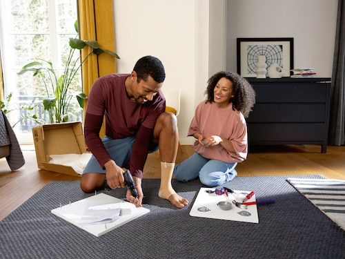 Smiling man sitting on the floor doing home repairs, wearing a beige Actimove Arthritis Care Ankle Support, getting help from partner
