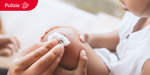 Etichetta per la pulizia con l'icona di una ferita su una mano sopra l'immagine di una madre che disinfetta la ferita al ginocchio di una bambina con garza e antisettico.