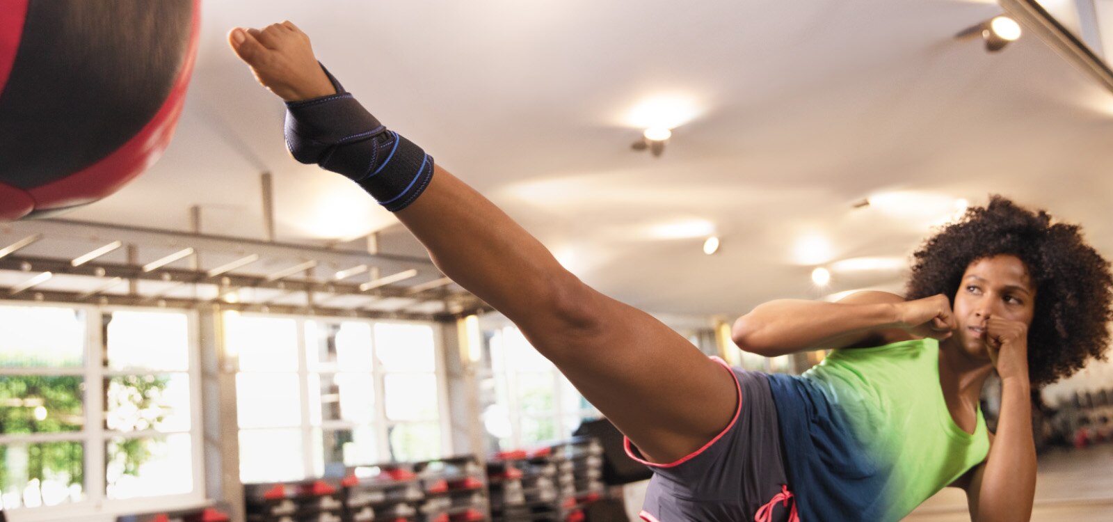 Kobieta kopiąca piłkę na siłowni podczas treningu z ortezą stawu skokowego Actimove Sports Edition z krzyżującymi się paskami