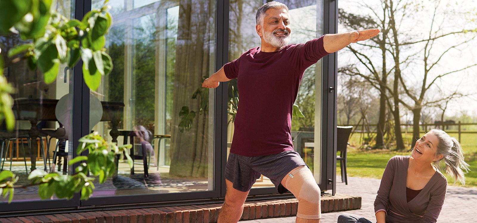 Smiling man wearing sports clothes and a knee support does stretch exercises outside as a woman looks on 