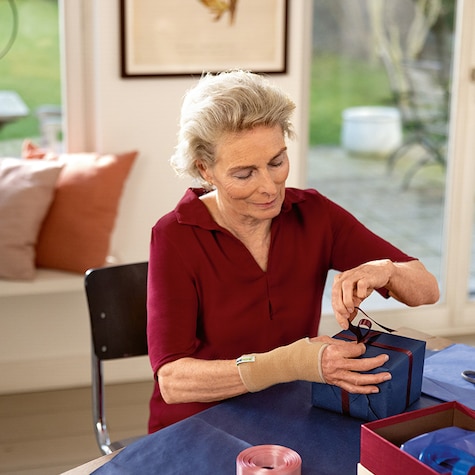 Older woman wearing a wrist support wraps ribbon around a wrapped gift on a table Older woman wearing a wrist support wraps ribbon around a wrapped gift on a table