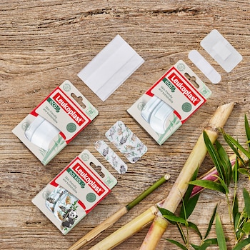 Overview of Leukoplast eco and Leukoplast eco kids dressings and packaging on a wooden background with bamboo sticks and leaves in the corner.