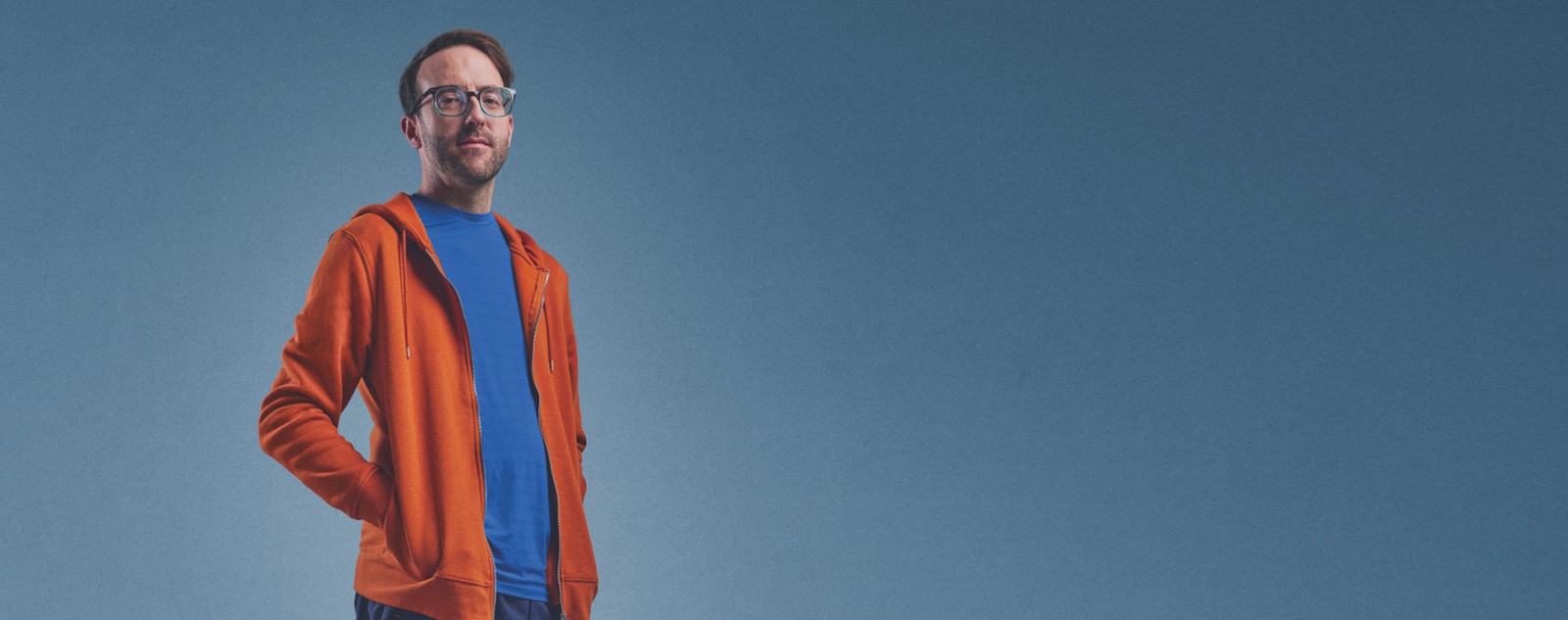 Andy står självsäkert med händerna i fickorna på sin orangea huvtröja.