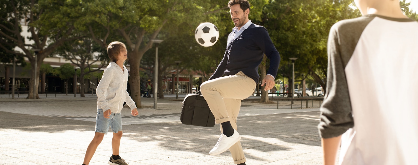 Ein Mann mit Kompressionsstrümpfen und Laptoptasche bleibt in einem Park stehen, um mit Kindern Fußball zu spielen.