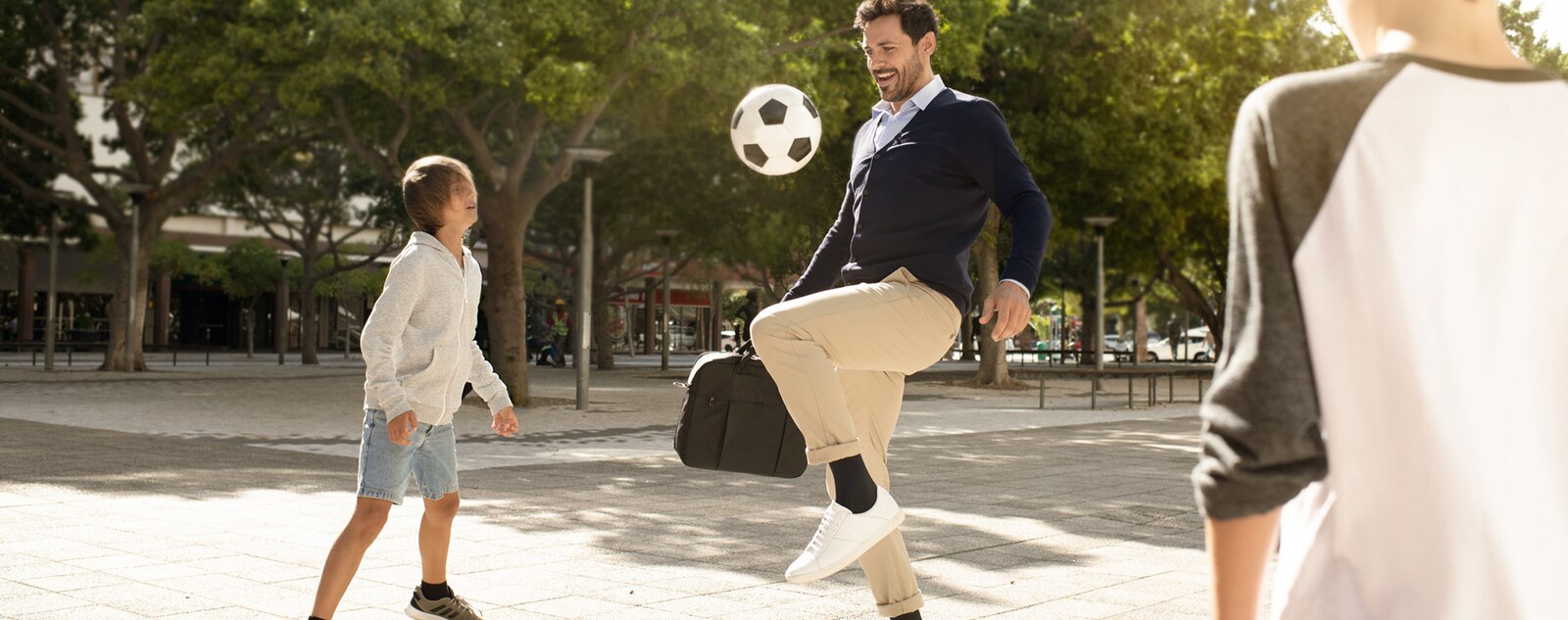 Man wearing compression stockings and carrying a laptop bag stops to play soccer with children in a park.