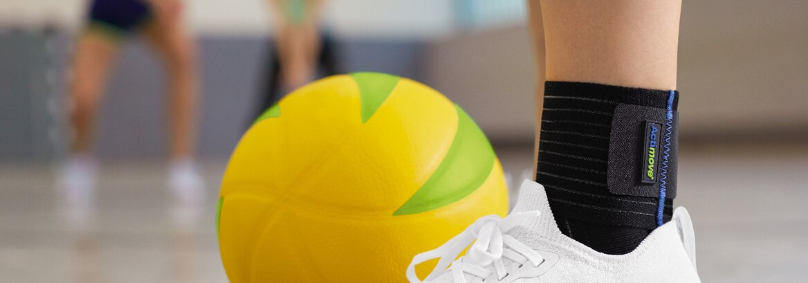 Noga w ortezie kostki i białych sznurowanych butach treningowych obok żółtej piłki