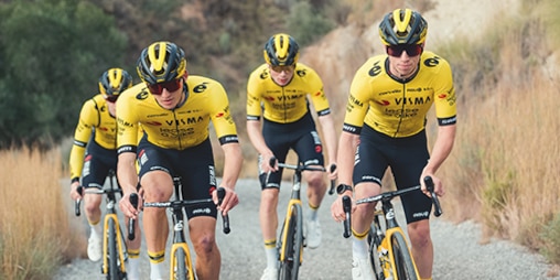 cykelryttere iført guletrøjer cykler ned ad en vej i et landligt landskab.