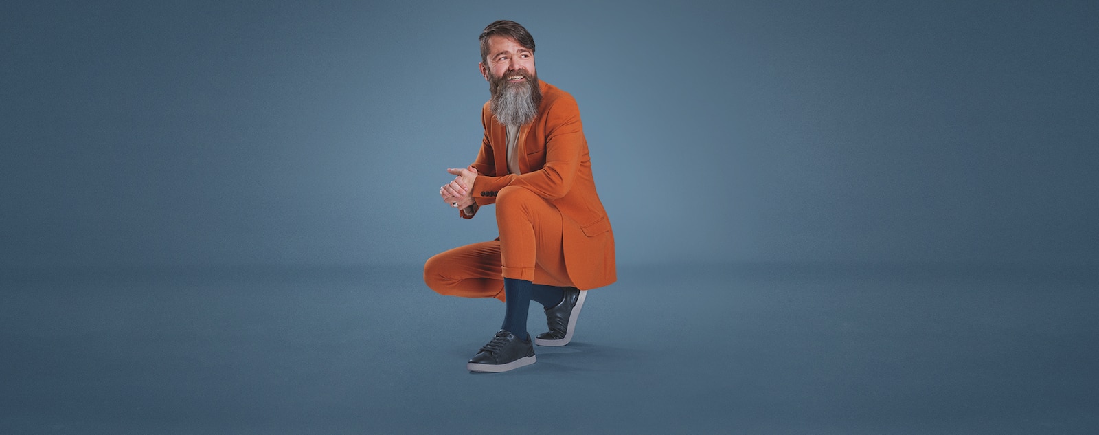 A man with a long beard, crouching down wearing a stylish orange suit and a pair of blue JOBST forMen socks