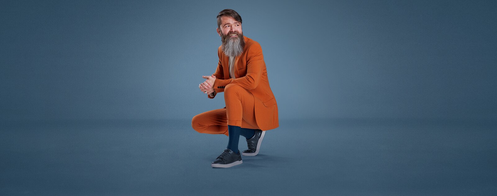 A man with a long beard, crouching down wearing a stylish orange suit and a pair of blue JOBST forMen socks