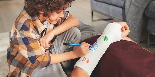 A young boy drawing on someone's arm cast with coloring pens.