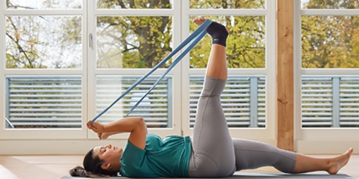 Woman wearing an ankle support lies down on the floor and uses a resistance band to stretch her leg