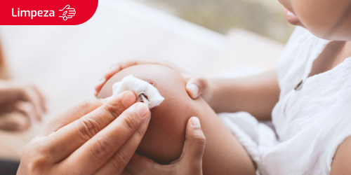 Etiqueta de limpeza com o ícone de uma ferida numa mão sobre uma imagem de uma mãe a desinfetar a ferida no joelho de uma menina com gaze e antissético.