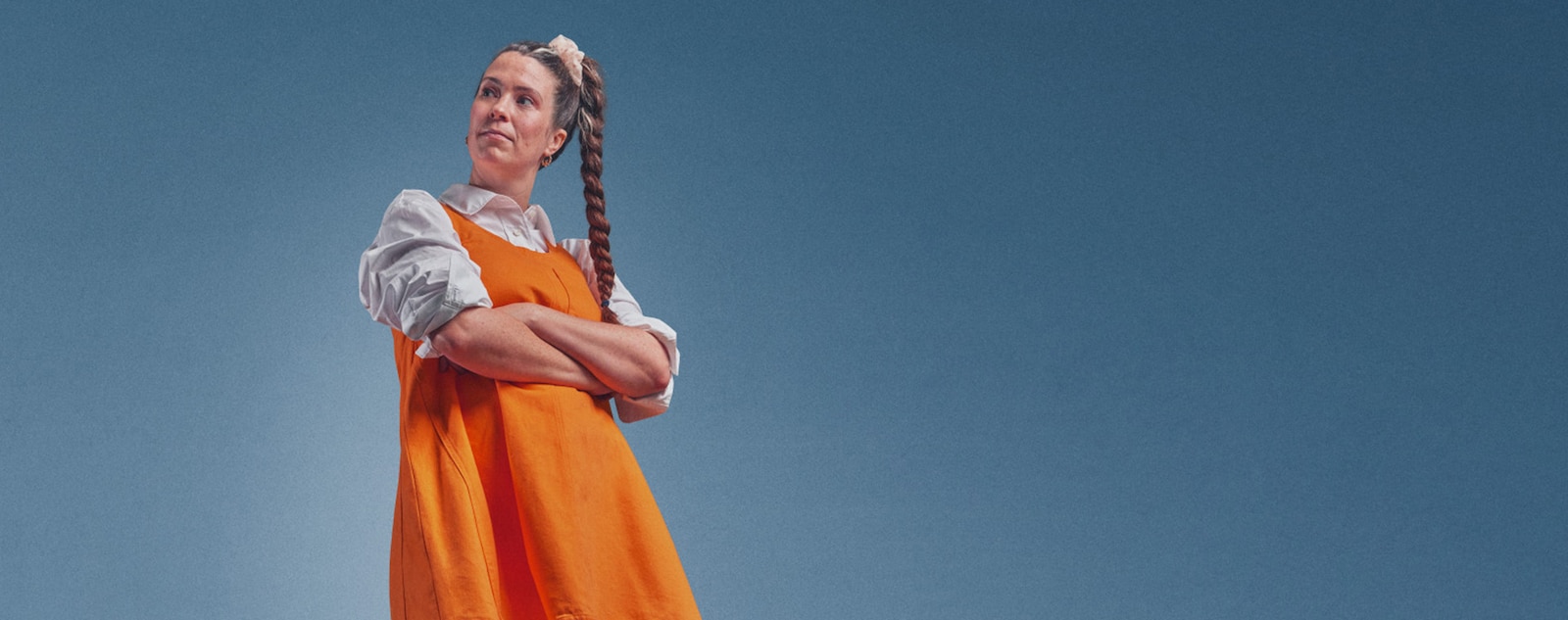 Kelley står med armarna i kors och bär en orange klänning över en vit skjorta.