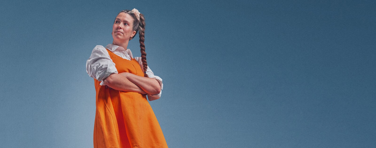 Kelley standing with arms crossed, wearing an orange dress over a white shirt.