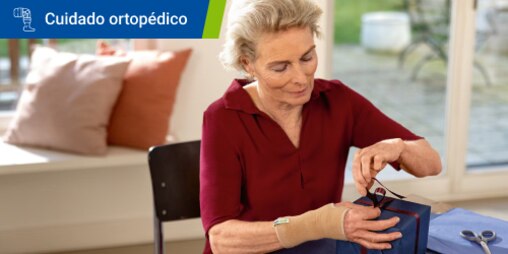 Una mujer mayor con una muñequera envuelve un regalo con una cinta sobre una mesa