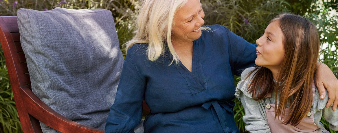 A woman wearing a blue short is holding her arm around a young girl while sitting outside in a garden