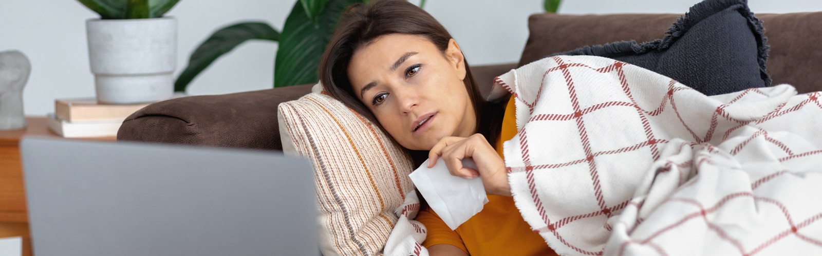 Eine Frau liegt krank auf der Couch und schaut in ihren Laptop