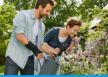 Mężczyzna noszący opaskę na nadgarstek trzyma konewkę, podlewając ogród, podczas gdy kobieta obok niego wącha kwiaty