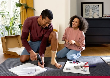 Man doing DIY and wearing an ankle support drills nails into the back of a square board while a woman looks on