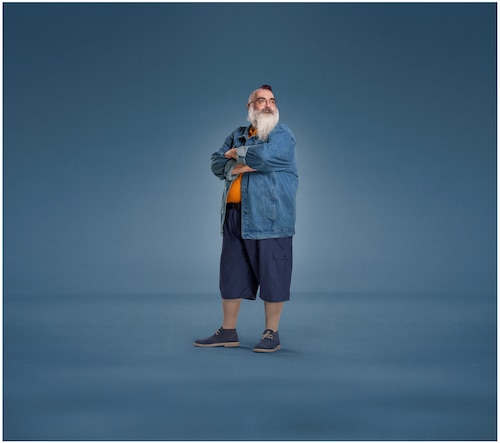 A bearded man with crossed arms, dressed in a denim jacket, orange shirt, navy shorts, and sneakers, wearing JOBST UlcerCare compression socks.