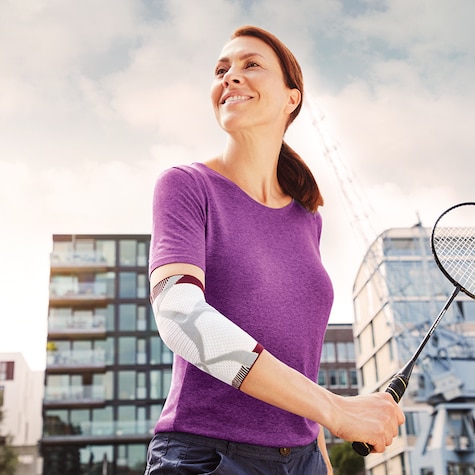 Kvinne iført en lilla topp og albuestøtte smiler og holder på en badmintonracket Kvinne iført en lilla topp og albuestøtte smiler og holder på en badmintonracket