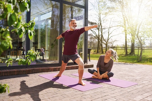 Smiling man wearing Actimove Arthritis Care Knee Support, practicing yoga outdoors with partner
