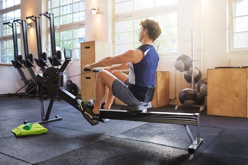 Man at gym working out at rowing machine, wearing an Actimove Sports Edition Back Stabilizer with Pressure Pads
