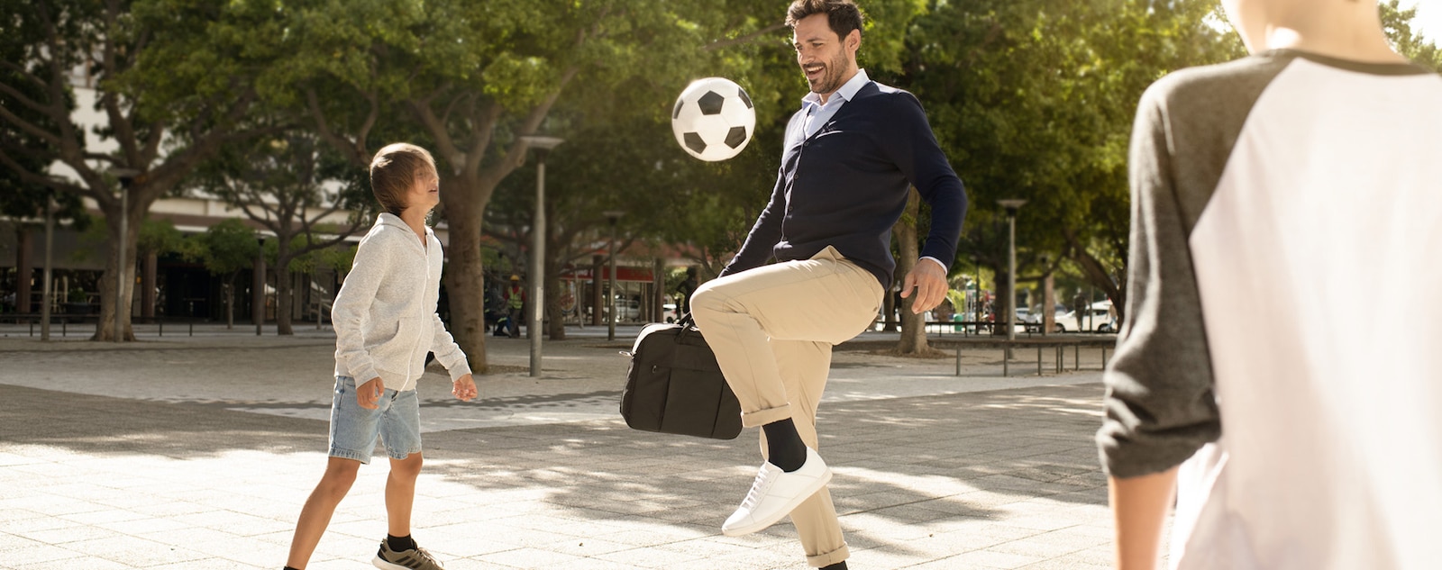 Man wearing compression stockings and carrying a laptop bag stops to play soccer with children in a park.