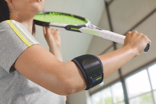 Primer plano de una mujer jugando a tenis en un gimnasio cubierto, usando una cinta para el codo Actimove Sports Edition con bolsa de frío/calor. 
