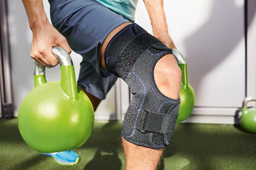 Close up of man lifting weights, wearing Actimove Sports Edition Knee Brace Wrap Around with Simple Hinges and Condyle Pads
