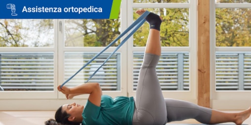 Una donna con una cavigliera distesa a terra, mentre usa una fascia elastica per lo stretching delle gambe