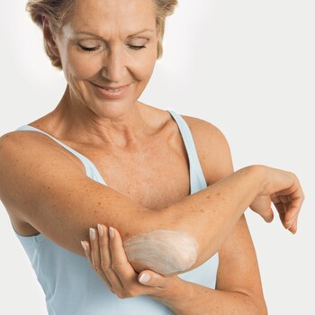 Une femme âgée applique Cutimed ACUTE Crème moussante sur son coude, une zone cutanée réputée sèche.
