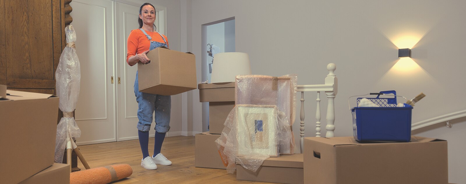 A person standing in a living room wearing casual clothes and compression stockings while holding a big box