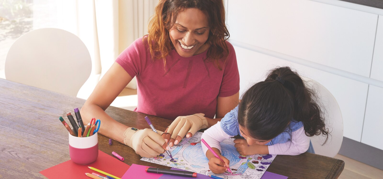 Mujer y niña coloreando un dibujo. La mujer lleva puestos los guantes para artritis Actimove Arthritis Care porque tiene molestias en el pulgar.
